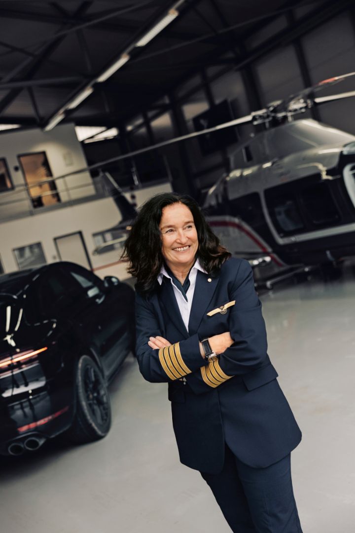A female pilot smiles while standing with arms crossed inside a hangar with a helicopter and car in the background.