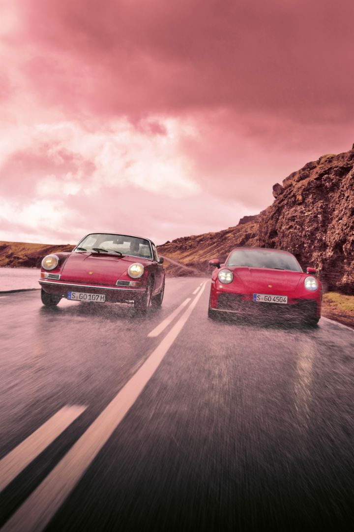 Two Porsche cars drive side by side along a wet road by a rocky cliff with a dramatic, stormy sky above. 