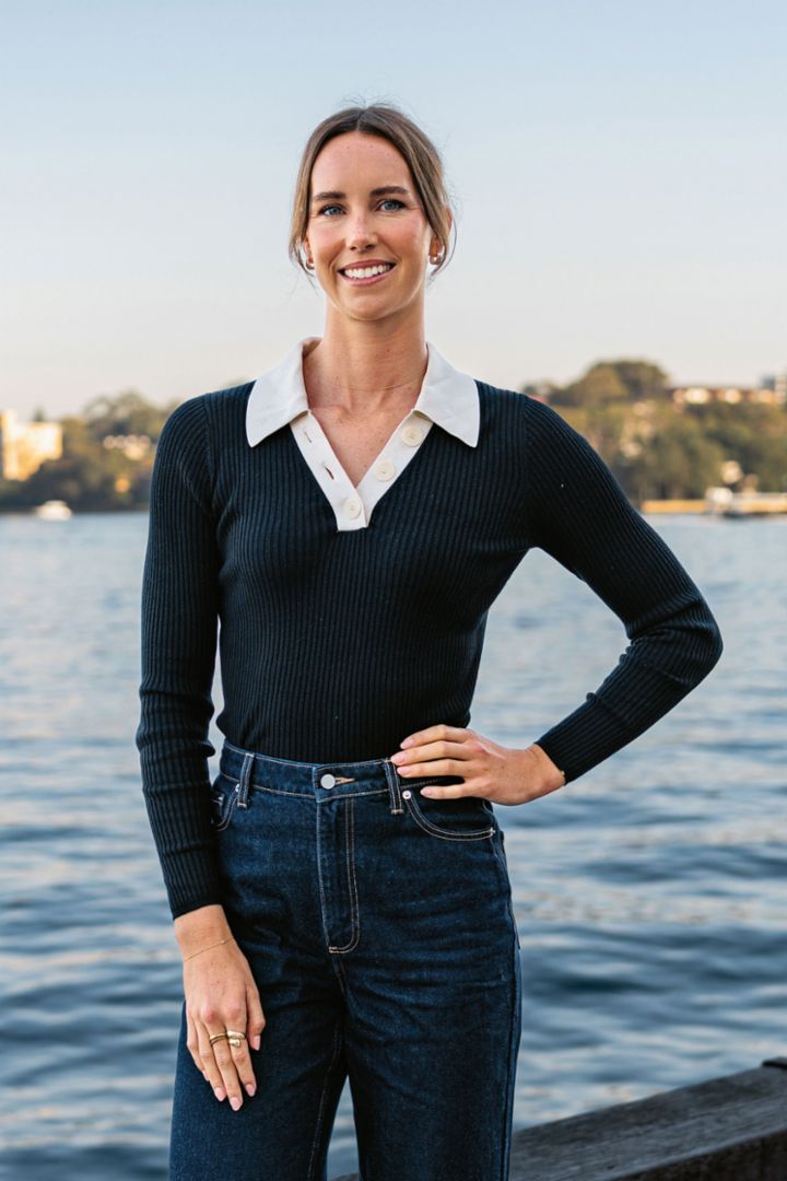 A woman wearing dark clothing stands by the water—there is an island in the background. 