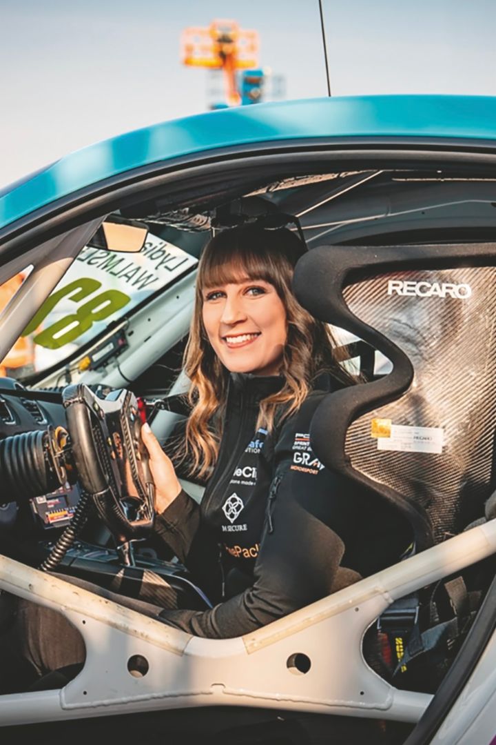 A woman in a racing suit sits in a race car, holding the steering wheel and smiling at the camera.