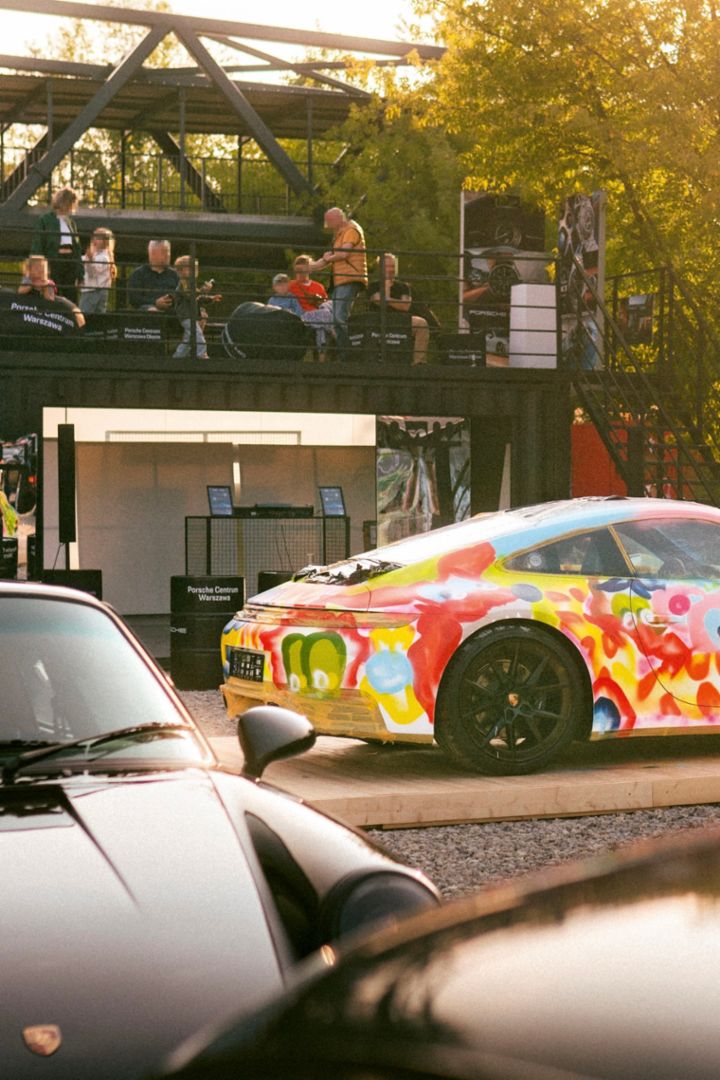 A Porsche 911 Turbo is parked on a driveway in front of a white modern building with large windows and palm trees in the background.
