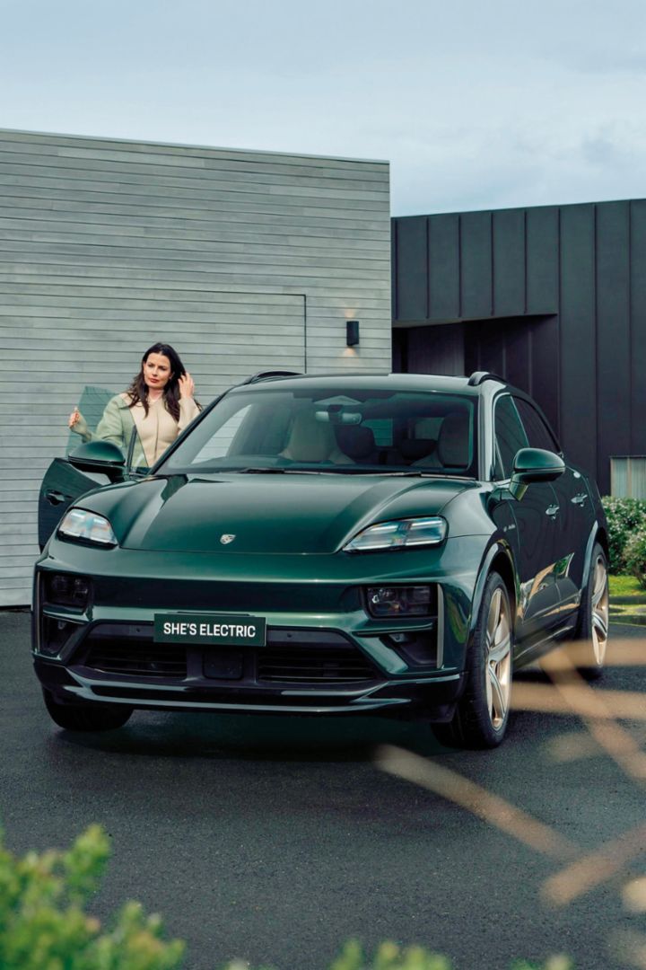 A woman with dark hair stands by the open driver's door of a dark Porsche SUV.