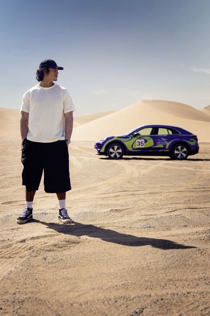 Ein Mann mit weißem T-Shirt und schwarzen Shorts steht auf einer sandigen Wüste, neben ihm steht ein bunter Porsche Macan.