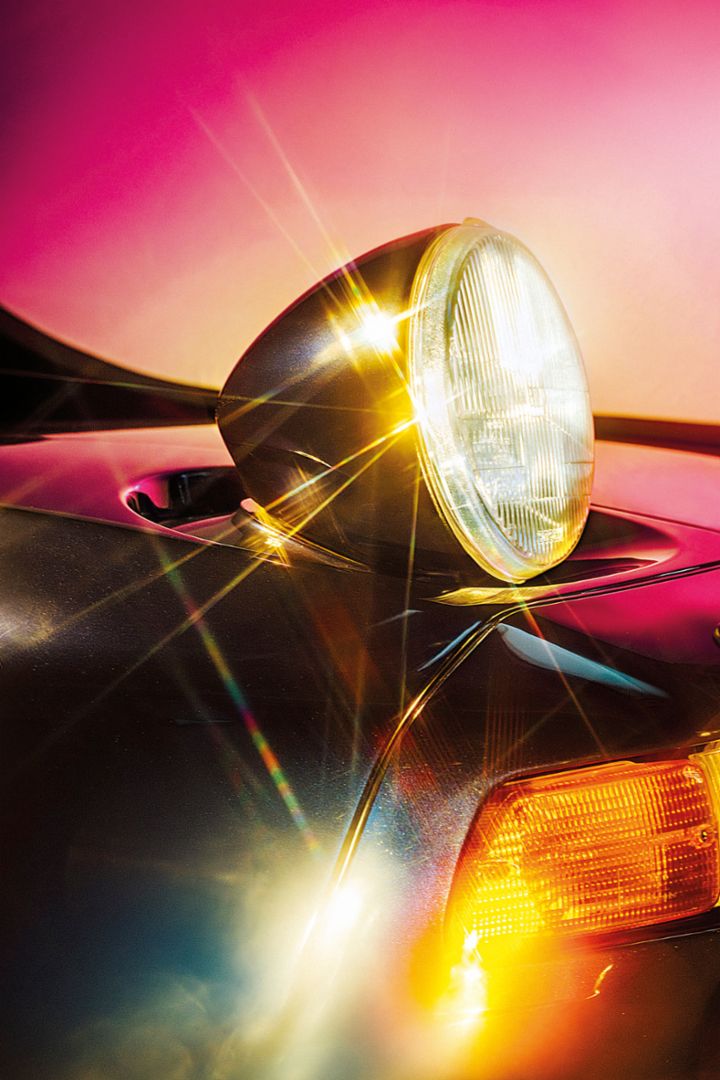 A woman stands smiling next to a Porsche 924, with the sun shining in the blue sky and the surroundings bathed in sunlight
