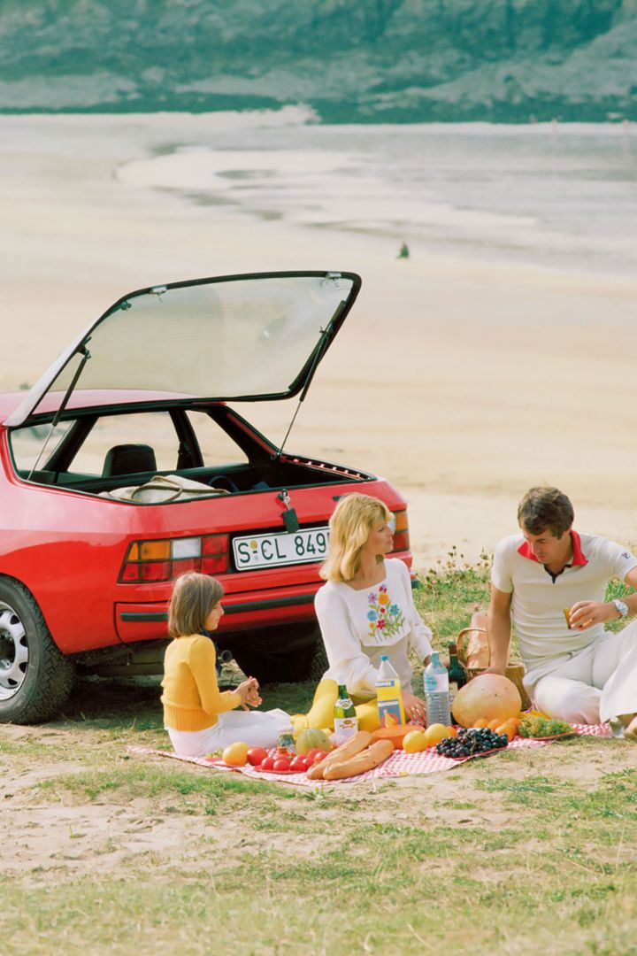 A family enjoys a picnic in front of a red Porsche 924 on the beach, with a wide view of the sea in the background.