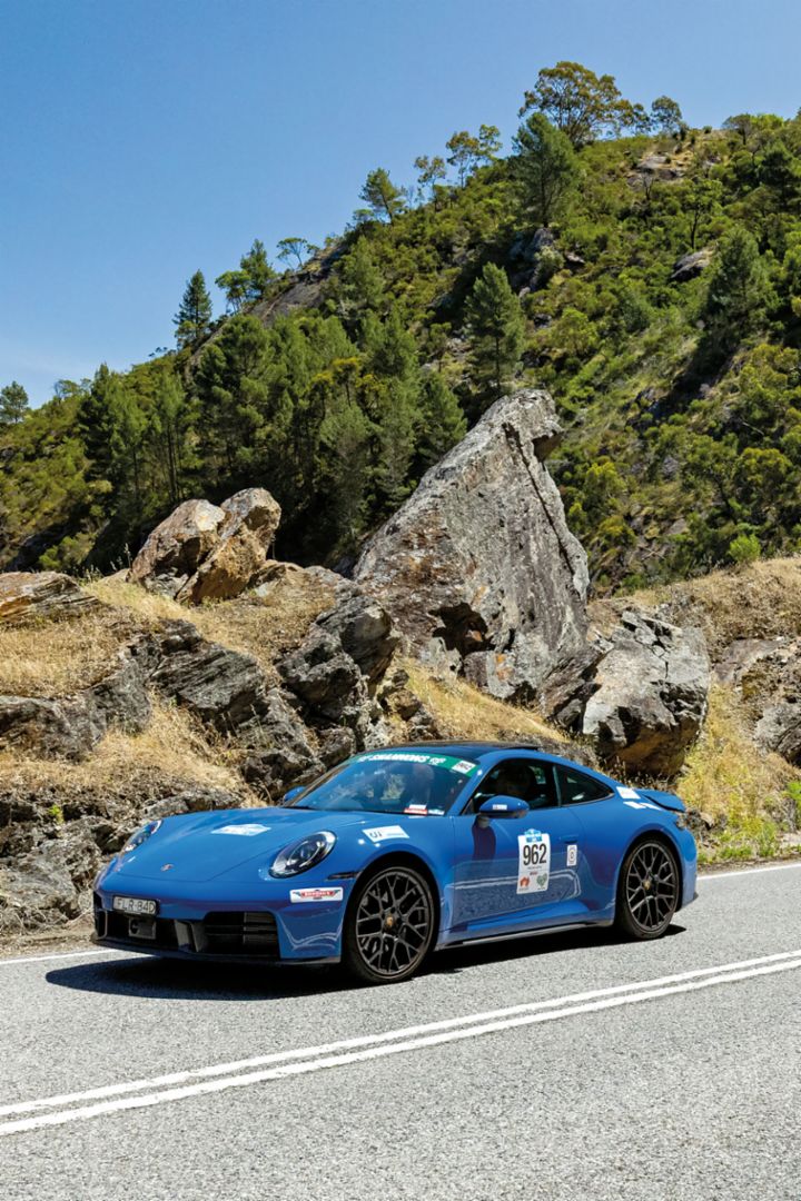 A blue Porsche 911 drives on a winding road through a mountainous landscape. The road is surrounded by rocks and lush greenery, under a clear blue sky.