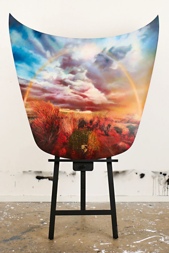 A large artwork painted on a car hood is displayed on an easel in a studio.