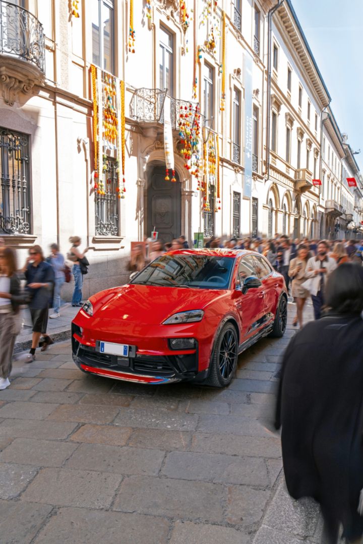 A woman in a purple dress is sitting inside a red cabriolet and holds the hand of a man who is standing outside of the car.