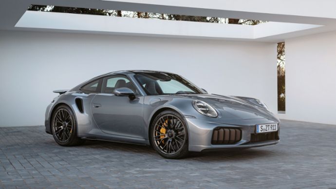 A Porsche 911 Turbo is parked on a driveway in front of a white modern building with large windows and palm trees in the background.
