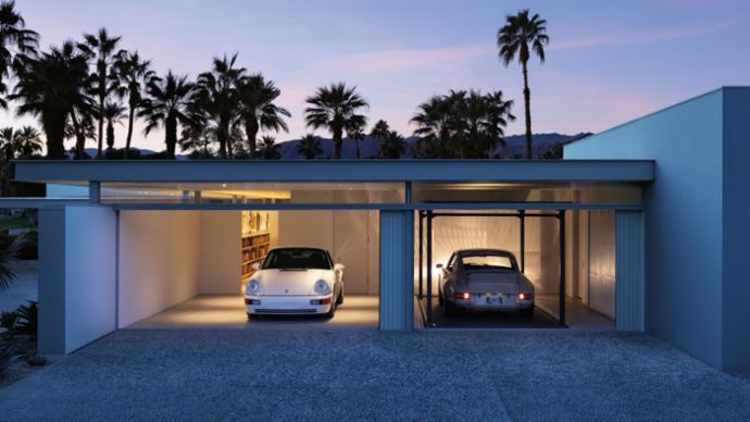 Two classic Porsches stand in a modern, illuminated garage building, surrounded by palm trees and at dusk.