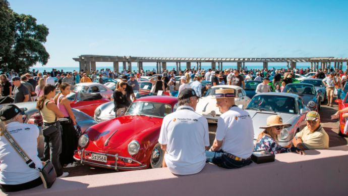 A group of people are looking at sports cars from different eras parked by the sea. 