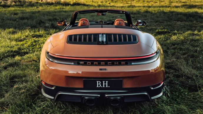 A brown Porsche 911 with the letters B. H. on the license plate is parked on a green lawn.