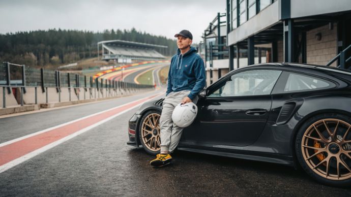 A man in casual clothes leans on a black Porsche 911 Turbo in front of a racetrack, holding a helmet in his hand.