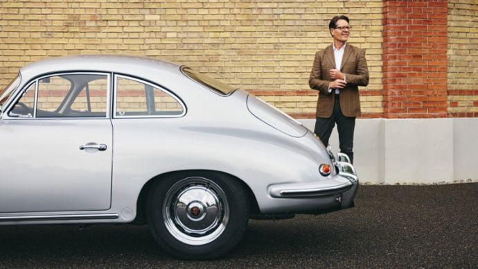 Ein Mann in einem braunen Blazer steht neben einem silbernen Porsche 356 und lächelt vor einer Backsteinwand.