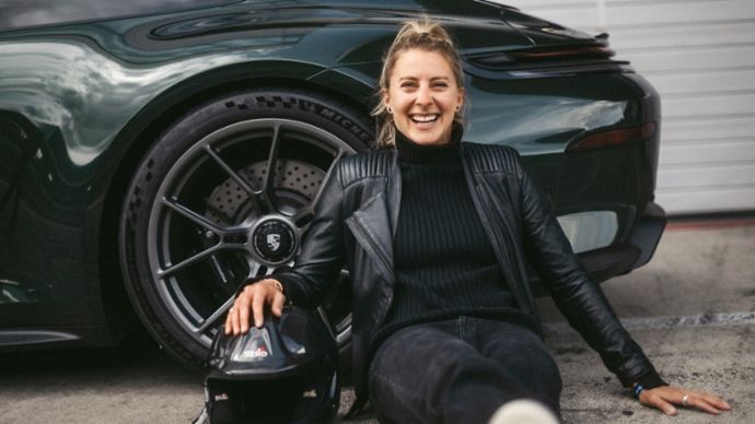Eine lachende Frau sitzt auf dem Boden neben einem grünen Porsche 911 und hat ihre Hand auf einem Helm abgelegt. 