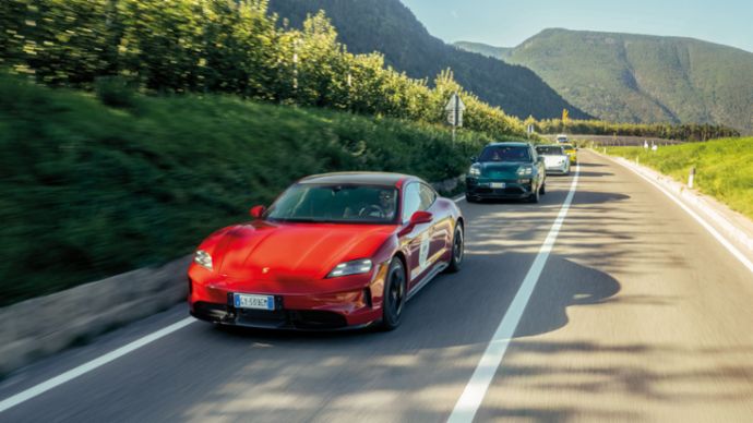 A red Porsche Taycan leads a convoy of cars driving along a country road through a mountainous landscape.