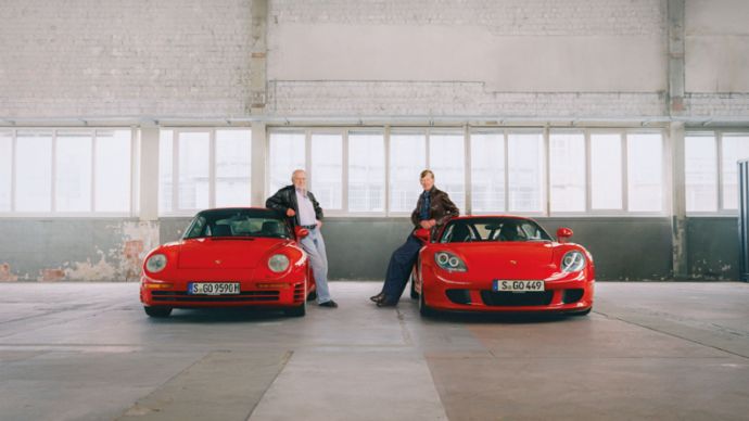 Two men lean against red Porsche sports cars in an industrial hall, parked side by side.