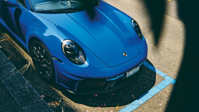 A blue Porsche sports car is parked in a marked spot, partially shaded with a view of the car’s front.