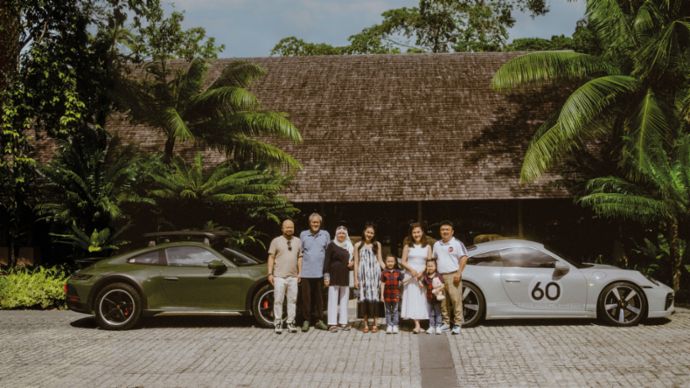 A group of people standing in a sunny setting, with two Porsche cars (green and silver) in the background in front of a tropical building.