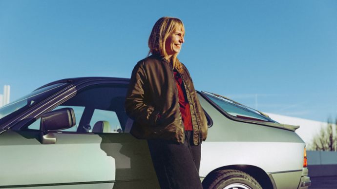 A woman stands smiling next to a Porsche 924, with the sun shining in the blue sky and the surroundings bathed in sunlight