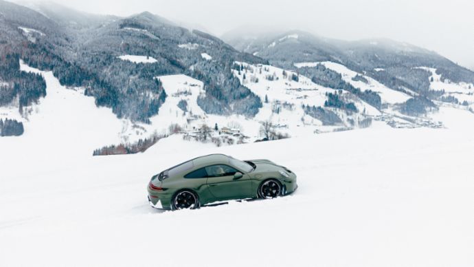A Porsche 911 drives through the snow, with a winter mountain landscape in the background.