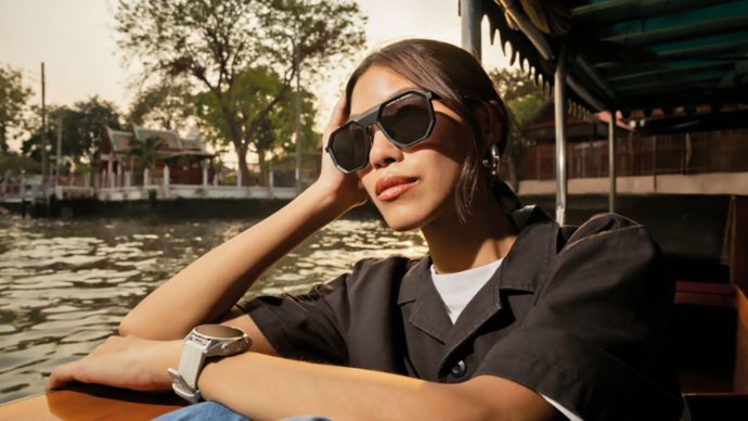 A young woman sits relaxed in a boat, wearing black Porsche Design sunglasses and a black jacket, looking into the distance. 