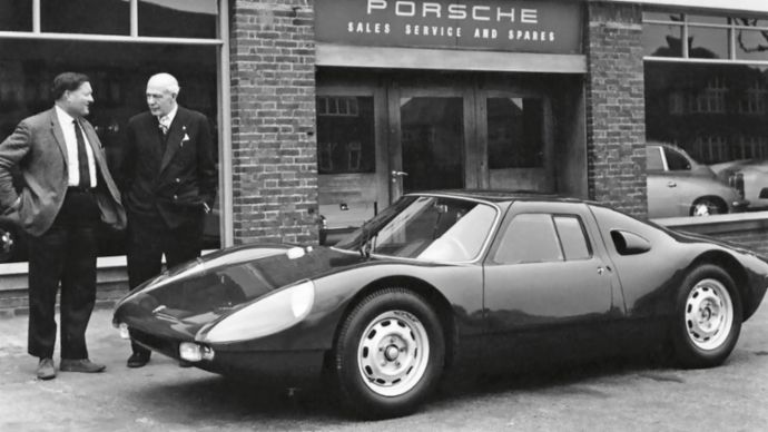 A black-and-white photo of a Porsche 904 in front of a Frazer Nash Cars dealership, with two men standing beside the car, capturing a historic moment in Porsche's early days in the UK.