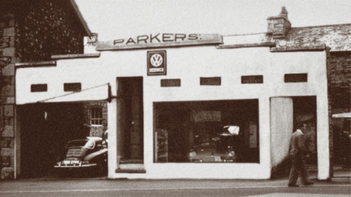 A historic photograph shows a small car dealership with vehicles outside and inside the showroom.