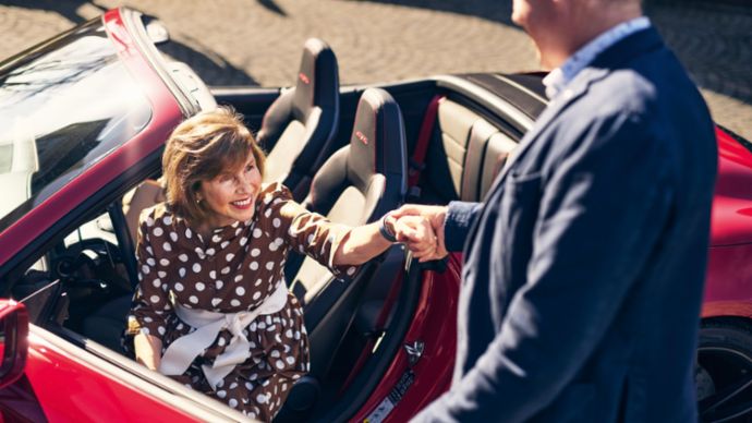 Eine Frau in einem gepunkteten Kleid sitzt in einem roten Cabriolet und hält die Hand eines Mannes, der außerhalb des Autos steht.