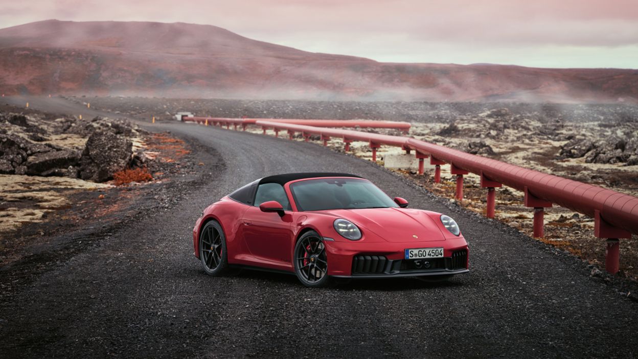 Ein roter Porsche 911 ist auf einer gewundenen, staubigen Straße geparkt, umgeben von vulkanischem Gelände und einem klaren Himmel darüber.