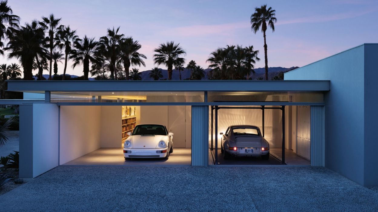 Zwei klassische Porsche stehen in einem modernen, beleuchteten Garagengebäude, umgeben von Palmen und bei Dämmerung.