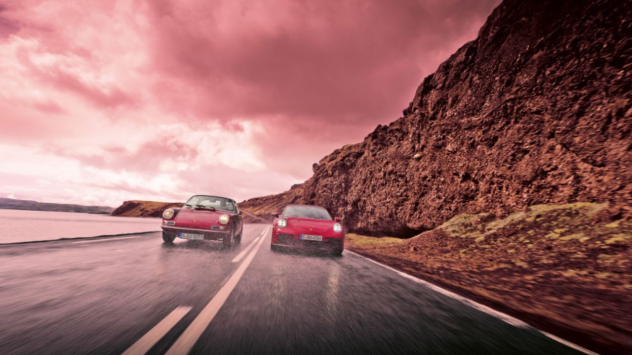 Zwei Porsche-Fahrzeuge fahren nebeneinander auf einer nassen Straße an einer felsigen Klippe entlang – darüber ein dramatischer, stürmischer Himmel.