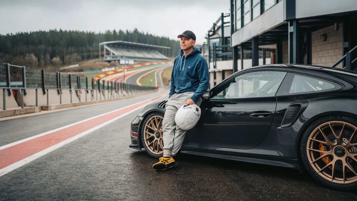 A man in casual clothes leans on a black Porsche 911 Turbo in front of a racetrack, holding a helmet in his hand.