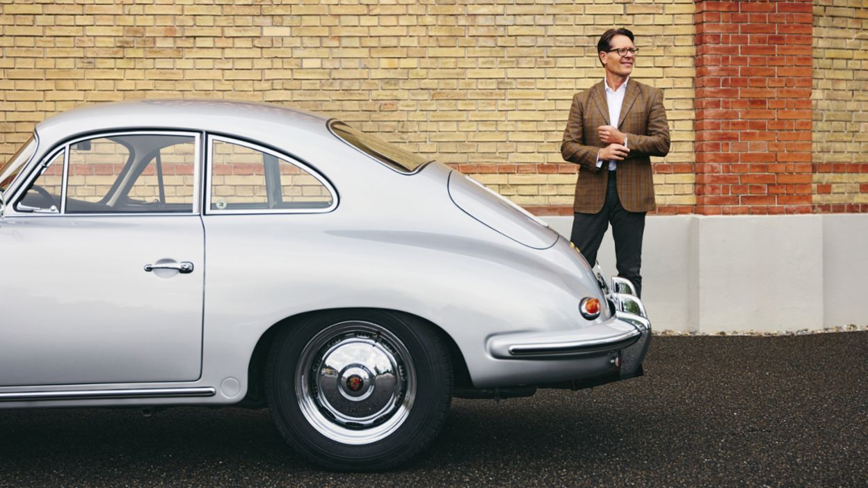 Ein Mann in einem braunen Blazer steht neben einem silbernen Porsche 356 und lächelt vor einer Backsteinwand.