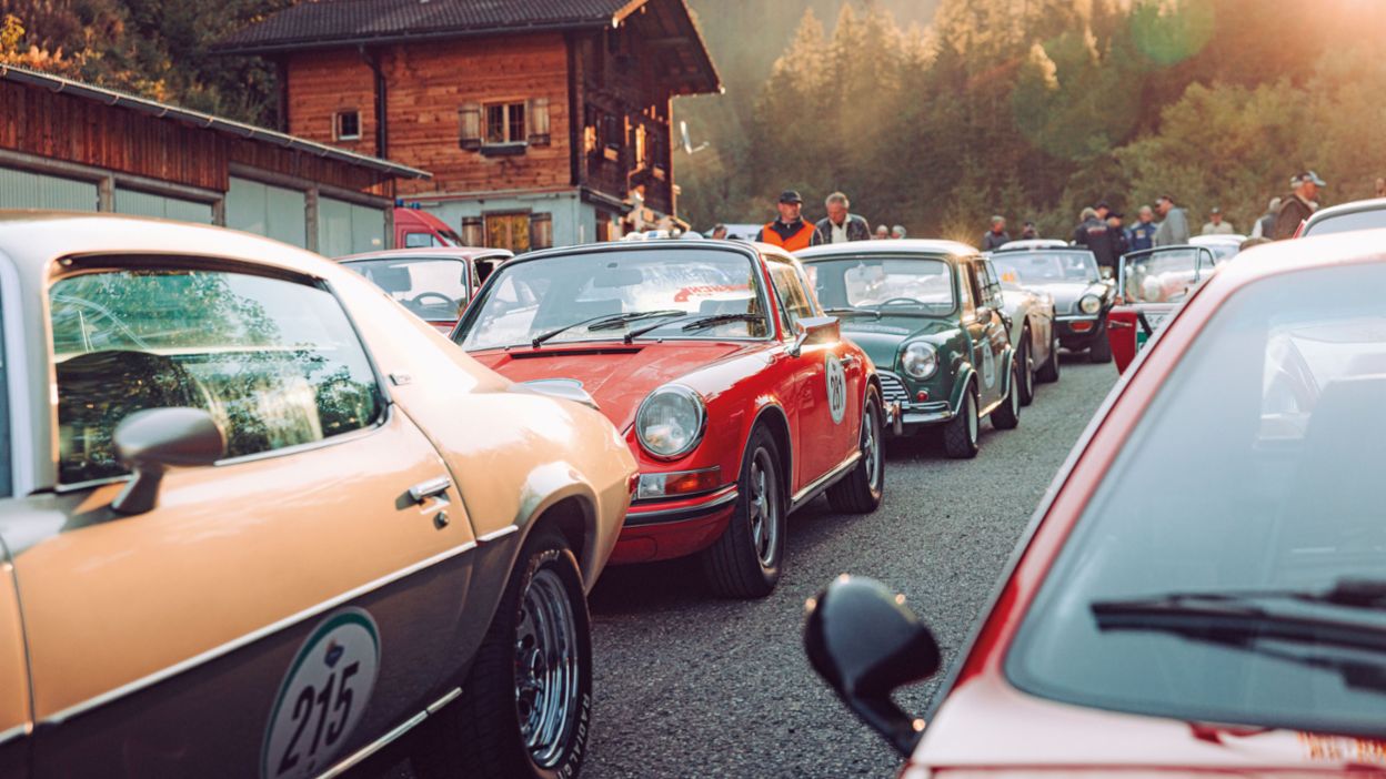 Mehrere klassische Rallye-Fahrzeuge stehen dicht aneinandergereiht vor einem alpinen Holzhaus, während Menschen sich zwischen den Autos versammeln.