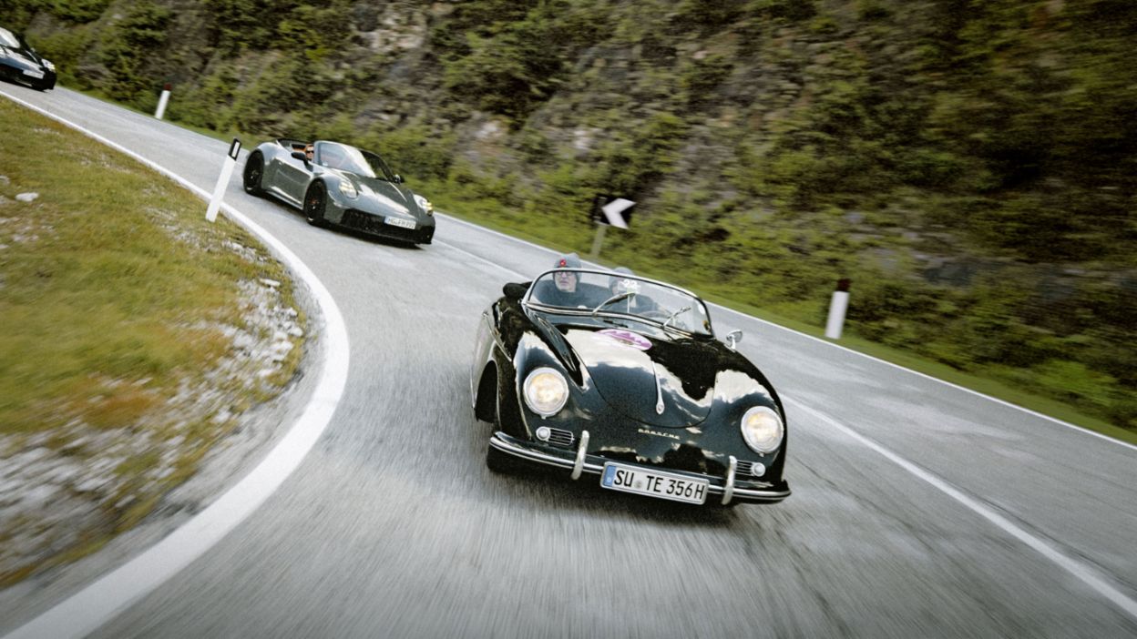 Zwei Porsche Cabriolets fahren auf einer kurvenreichen Straße in einer malerischen Berglandschaft mit grünen Bäumen zu beiden Seiten.