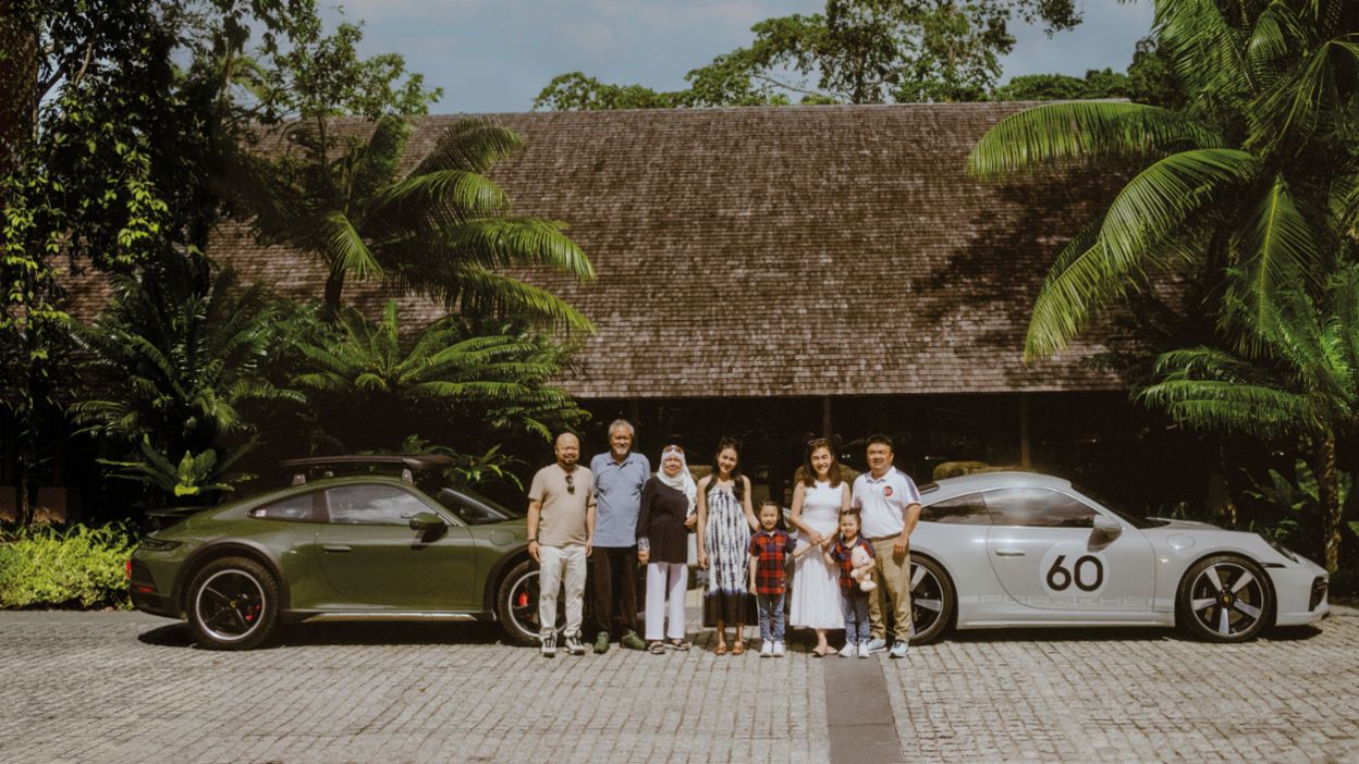 A group of people standing in a sunny setting, with two Porsche cars (green and silver) in the background in front of a tropical building.