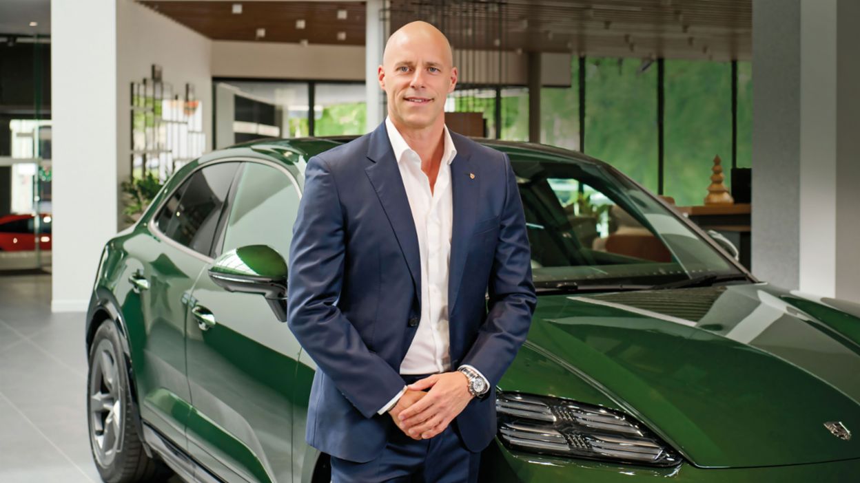 A man with his hands folded and wearing a business stands next to a green Porsche Macan in a showroom. He is smiling and wearing a white shirt, with his hands folded in front of him. The showroom has modern decor, with soft lighting and a view of a green outdoor space through the windows.