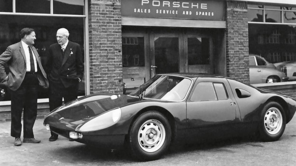 A black-and-white photo of a Porsche 904 in front of a Frazer Nash Cars dealership, with two men standing beside the car, capturing a historic moment in Porsche's early days in the UK.