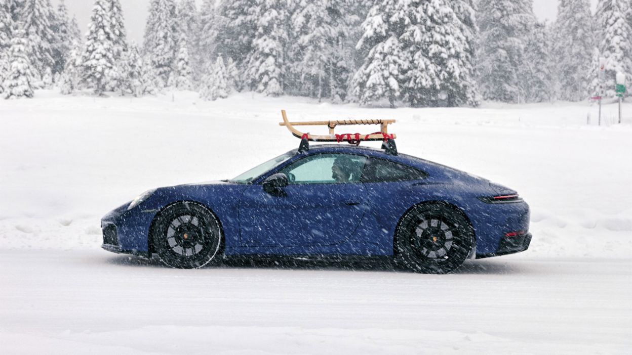 A blue Porsche 911 driving through a snowy landscape, with a ski rack on the roof, surrounded by dense, snow-covered trees. 